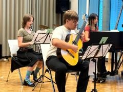 Drei Jugendliche musizieren gemeinsam. Ein Junge spielt vorne Gitarre. Dahinter sitzt ein Mädchen am Klavier. Neben ihr sitzt ein Mädchen und spielt Klarinette.