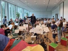 Ein Mann dirigiert ein Kinderorchester. Hinter dem Orchester sitzten viele Zuhörer:innen.