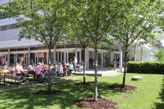 Musikschule von außen im Sommer. Einige Menschen sitzen auf Bierbänken in der Sonne.