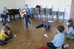 Eine Gruppe von Kindern sitzt im Kreis. Ein Musikschullehrer spielt Gitarre.