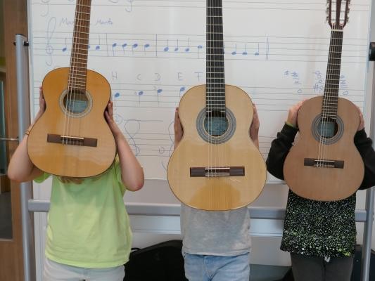 Drei Kinder stehen vor einer Tafel mit Musiknoten und halten Gitarren hoch.