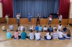 Kinder sitzen in eienr Turnhalle. Vor ihnen führen die Musikschullehrer verschiedene Instrumente vor.