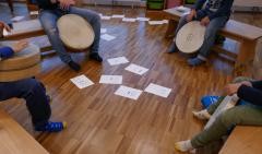Trommelgruppe: mehrer Kinder mit Trommeln sitzen in einem Kreis. Auf dem Boden liegen verschiedene Blätter.