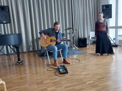 Eine singende Musiklehrerin wird von einem Gitarrenspielenden Lehrer begleitet.