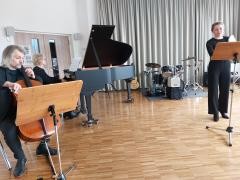 Eine singende Musiklehrerin wird von einem Cellospieler und einer Flügelspielerin begleitet.