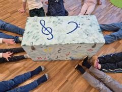 Viele Kinder sitzen um eine mit Noten und Notenschlüsseln bemalte Kiste.